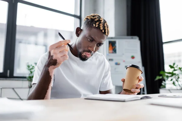 Hombre afroamericano serio con vitiligo sosteniendo taza de papel y pluma mientras mira en el cuaderno en la oficina - foto de stock