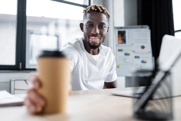 Young african american man with vitiligo skin smiling near blurred paper cup in office - foto de stock