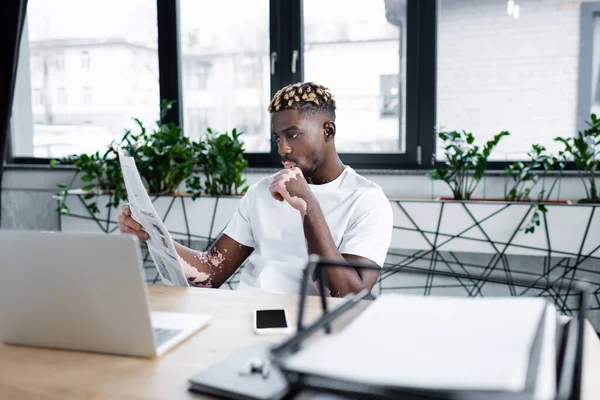 American man with vitiligo skin and trendy hairstyle reading newspaper near laptop and smartphone in office — стокове фото