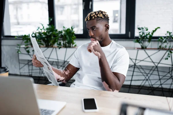 Hombre afroamericano de moda con periódico de lectura vitiligo cerca de computadora portátil borrosa y teléfono celular con pantalla en blanco - foto de stock