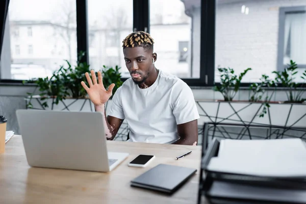 Trendy african american man with vitiligo waving hand during video call on laptop in office — Stock Photo