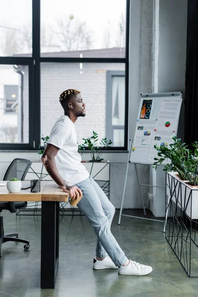 Side view of african american man with vitiligo skin standing in earphone near desk and blurred flip chart — Stock Photo