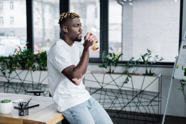 Joven afroamericano hombre con vitiligo beber café para ir mientras escucha música en auriculares en la oficina - foto de stock