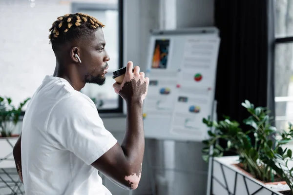 Vista lateral del hombre afroamericano con vitiligo bebiendo café y escuchando música en auriculares cerca del rotafolio borroso - foto de stock