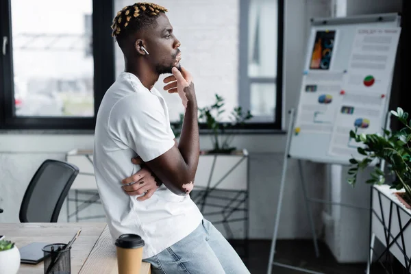 Side view of african american man with vitiligo listening music in earphone near coffee to go in office — Stockfoto