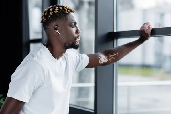 African american man with vitiligo listening music in earphone while standing with closed eyes near office windows — Fotografia de Stock