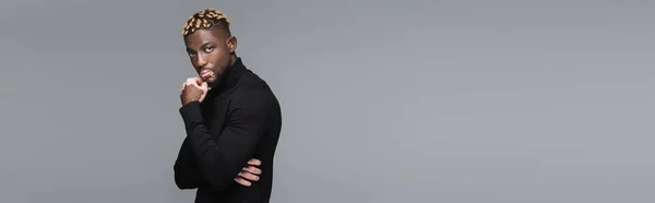 Hombre afroamericano de moda con vitiligo, con cuello alto negro, tocando la barbilla aislada en gris, pancarta - foto de stock