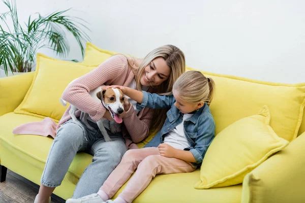 Smiling mother and daughter holding jack russell terrier on couch at home - foto de stock