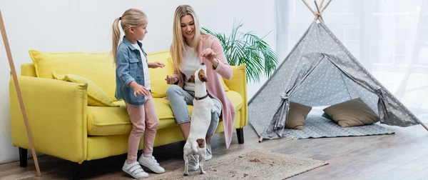 Smiling woman feeding jack russell terrier near kid at home, banner - foto de stock