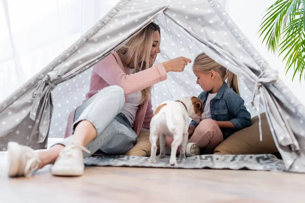 Positive mother and kid sitting in tent near jack russell terrier at home — Stock Photo