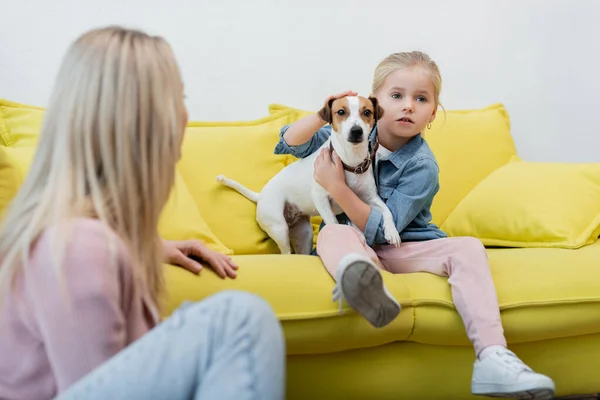 Kid hugging jack russell terrier near blurred mom at home — стоковое фото