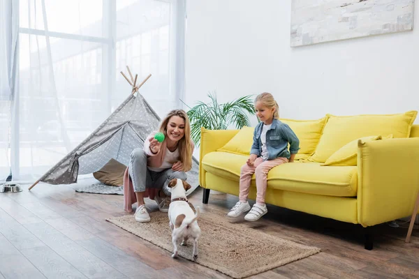 Smiling woman holding ball near jack russell terrier near daughter at home — Photo de stock