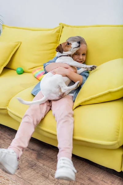 Kid looking at camera and hugging jack russell terrier on couch — Foto stock