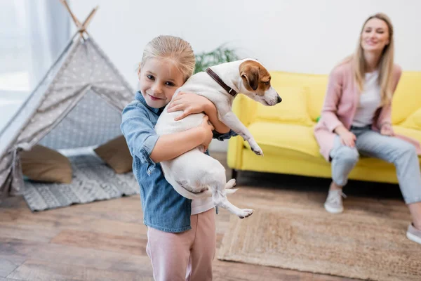 Happy kid hugging jack russell terrier near blurred mom at home - foto de stock