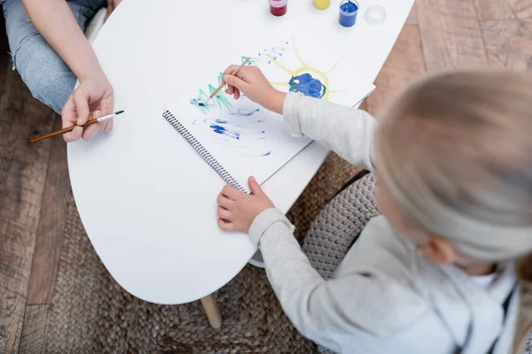 Overhead view of kid drawing on sketchbook near mom with paintbrush at home - foto de stock