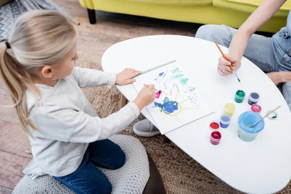 High angle view of blurred girl drawing on sketchbook near mom and gouache at home — Stock Photo