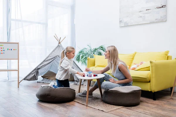 Side view of smiling mother and daughter drawing near couch and teepee at home — стоковое фото