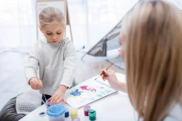 Daughter holding paintbrush near gouache and blurred mom at home — стоковое фото