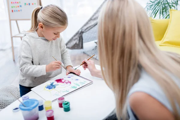 Kid drawing on sketchbook near gouache and blurred mom with paintbrush at home - foto de stock