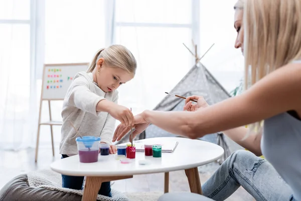 Blurred woman painting with kid near gouache and sketchbook at home — Stock Photo