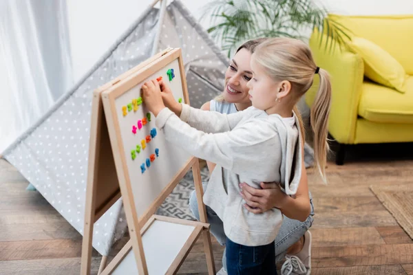 Smiling mother hugging kid near magnetic easel and blurred tent at home — стоковое фото