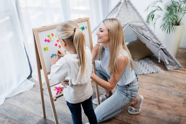 Woman looking at magnetic easel near daughter and magnets at home — Fotografia de Stock