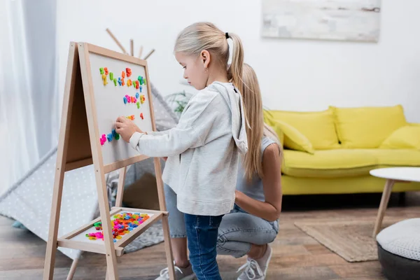 Girl playing with colorful magnets on easel near mom at home — Foto stock