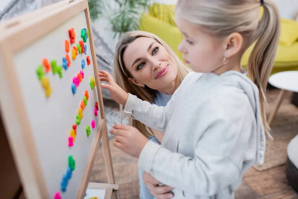 Smiling mother looking at daughter near easel with colorful magnets at home - foto de stock