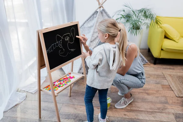 Child standing near mother with chalk and chalkboard at home — Stockfoto