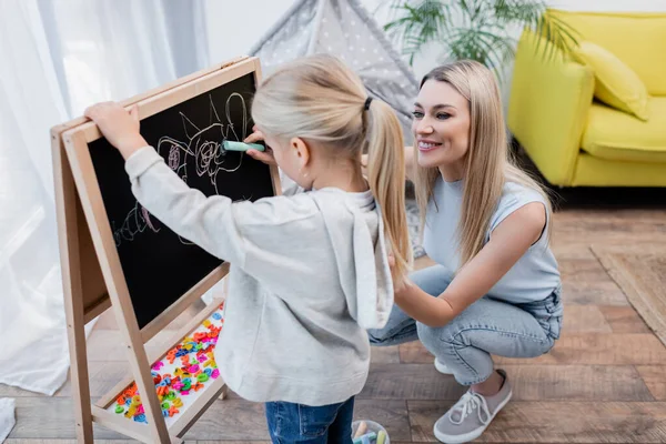 Positive woman looking at daughter drawing on chalkboard at home — Foto stock