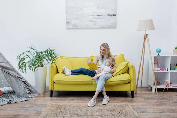 Positive woman reading book near daughter on couch at home — стоковое фото