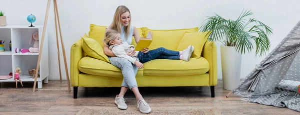 Woman reading book near kid on couch and teepee at home, banner - foto de stock