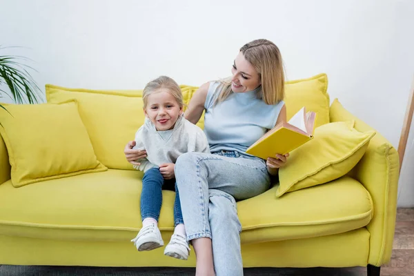 Kid sticking out tongue near mom with book on couch at home — Photo de stock