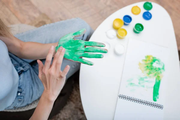 Top view of woman applying paint on hand near blurred sketchbook at home - foto de stock