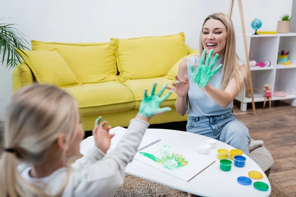 Smiling woman showing hand in paint near blurred daughter at home — Stock Photo