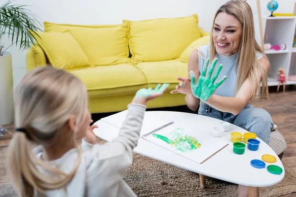 Positive mother showing hand in paint at blurred kid at home — стоковое фото