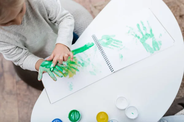 Top view of kid applying paint on hand near sketchbook on coffee table — Stockfoto