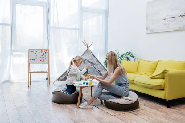 Side view of mom and kid applying paint on hands near sketchbook on coffee table at home — Photo de stock