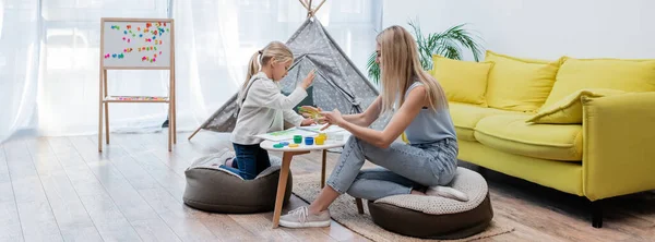 Side view of family applying paint on hand near sketchbook on coffee table and teepee at home, banner — Stockfoto