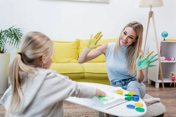 Smiling woman showing hands in paint near blurred kid at home — стоковое фото