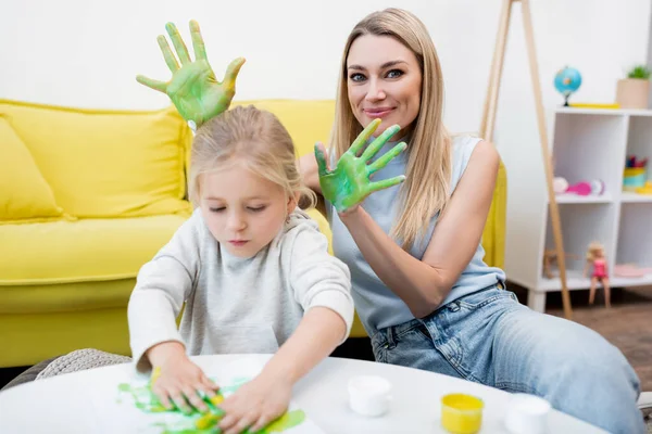 Woman holding hands in paint near kid drawing at home — стоковое фото