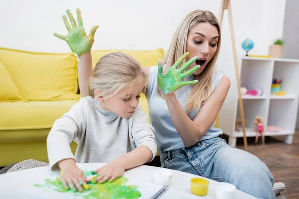 Shocked mother holding hands in paint near kid at home — стоковое фото