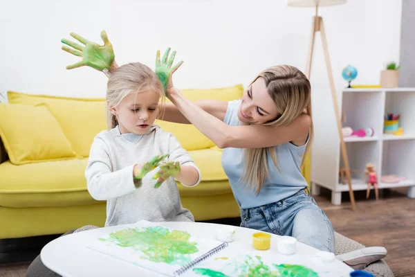 Positive woman holding hands in paint near daughter and sketchbook on coffee table at home — Stock Photo