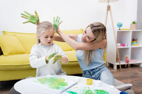 Positive woman holding hands in paint near daughter and sketchbook on coffee table at home — Stockfoto