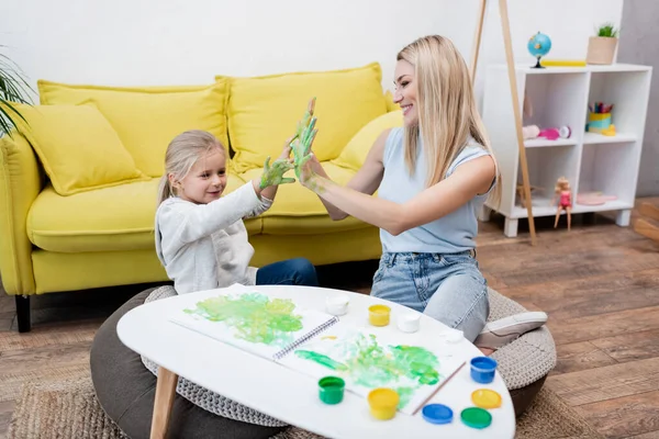 Smiling kid and mother showing hands in paint near sketchbook on coffee table in living room — стоковое фото