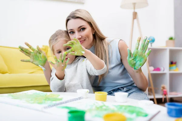 Woman looking at camera near daughter looking at hands in paint at home - foto de stock