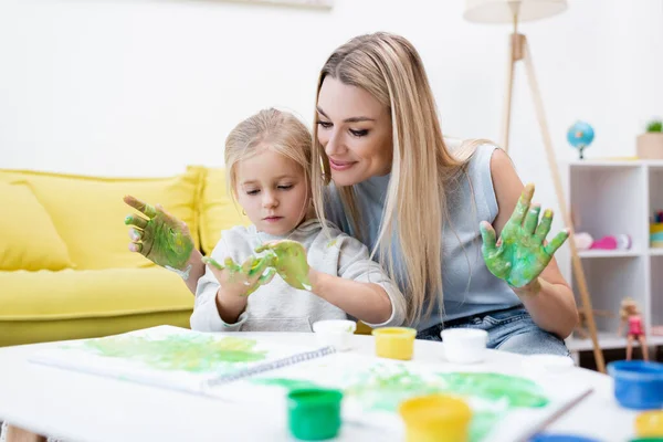 Woman looking at hands in paint near daughter at home - foto de stock