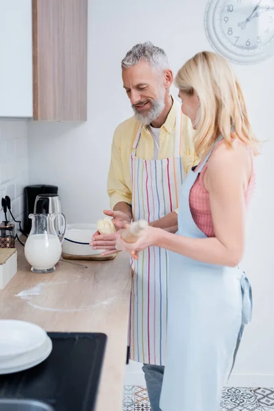 Smiling mature man in apron holding dough near wife with rolling pin in kitchen — Stock Photo