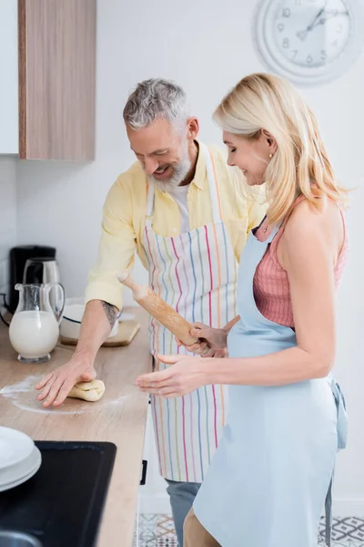 Couple mature positif tenant la pâte et le rouleau à pâtisserie dans la cuisine — Photo de stock