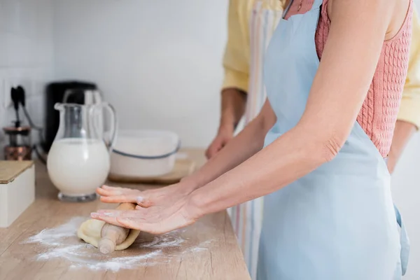 Обрезанный вид женщины в фартуке катит тесто со штифтом рядом с мужем на кухне — стоковое фото